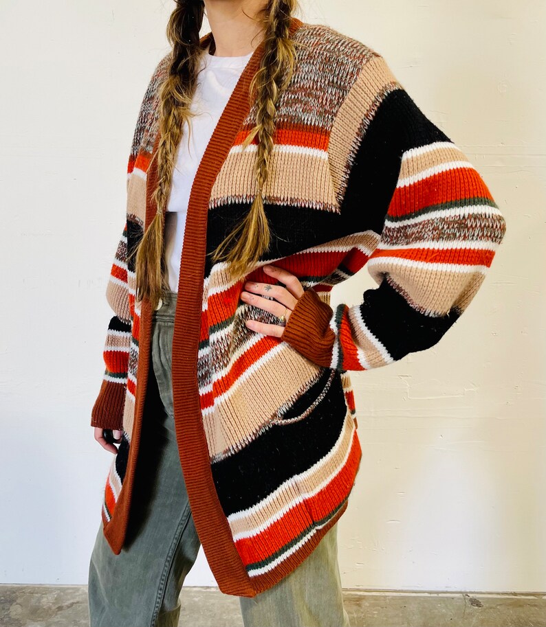 May include: A multi-colored open cardigan with a striped pattern in shades of brown, black, orange, and cream. The cardigan has long sleeves and a brown trim. The garment is made of a thick knit material.