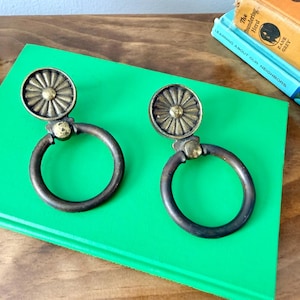May include: Two matching brass drawer pulls featuring a circular backplate with a sunburst or petal design and a hinged, rounded drop ring handle. The metal has a darkened, aged patina finish with visible texture on the circular mounting plates.