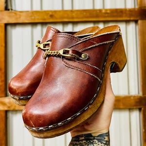 May include: Brown leather clogs with a wooden heel and a gold chain detail. The clogs have a white stitching detail around the sole.