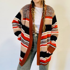 May include: A multi-colored open cardigan sweater with a striped pattern in shades of brown, black, orange, and white. The sweater has long sleeves and a relaxed fit. The cardigan is made of a soft, knitted material.