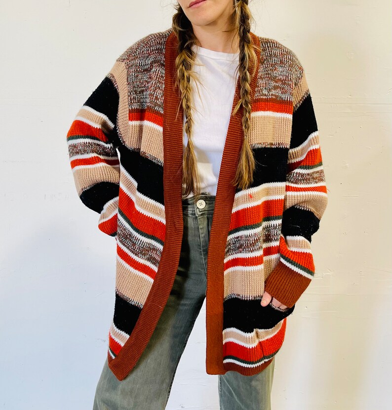 May include: A multi-colored open cardigan sweater with a striped pattern in shades of brown, black, orange, and white. The sweater has long sleeves and a relaxed fit. The cardigan is made of a soft, knitted material.