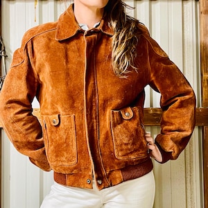 May include: A brown suede jacket with a collared neckline, two front pockets, and a ribbed hem. The jacket has a button closure and a classic bomber style. The sleeves are long and the jacket appears to be a medium size.