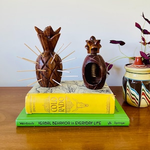 May include: Two wooden pineapple-shaped objects, one with toothpicks, sit atop books. The books are yellow and green, with the titles "Colorado" and "Verbal Behavior in Everyday Life." A decorative jar with a colorful design is in the background.