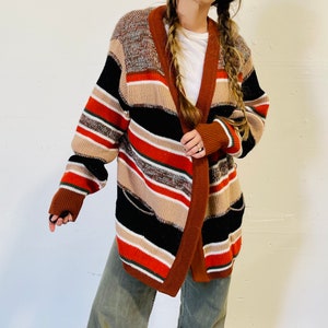 May include: A multi-colored striped cardigan in shades of brown, black, tan, and red. The open-front sweater has a shawl collar and pockets. The sleeves are long, and the cardigan appears to be made of a thick knit material.