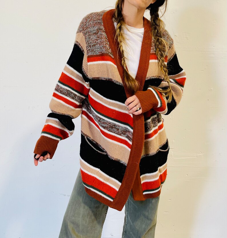 May include: A multi-colored striped cardigan in shades of brown, black, tan, and red. The open-front sweater has a shawl collar and pockets. The sleeves are long, and the cardigan appears to be made of a thick knit material.