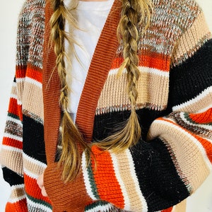 May include: A multi-colored, open-front cardigan sweater with a striped pattern in shades of brown, black, orange, and beige. The sweater has a wide, solid brown trim along the edges and sleeves. The sleeves are long and the sweater appears to be made of a knitted material.