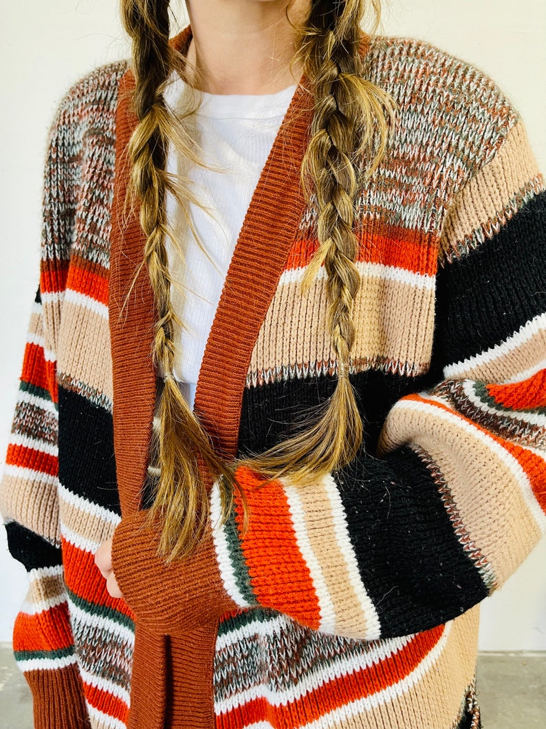 May include: A multi-colored, open-front cardigan sweater with a striped pattern in shades of brown, black, orange, and beige. The sweater has a wide, solid brown trim along the edges and sleeves. The sleeves are long and the sweater appears to be made of a knitted material.