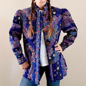 May include: A long-sleeved quilted jacket in shades of deep blue, purple, and magenta, featuring a patchwork design of floral and paisley patterns. The jacket has a high collar, puffed shoulders, and a front button closure with square buttons.