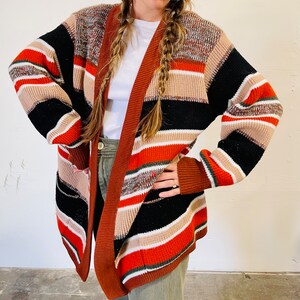 May include: A multi-colored, open-front cardigan sweater. The sweater features horizontal stripes in shades of black, orange, brown, beige, and white. The sleeves are long, and the sweater appears to be made of a knitted material.