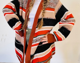 May include: A multi-colored, open-front cardigan sweater. The sweater features horizontal stripes in shades of black, orange, brown, beige, and white. The sleeves are long, and the sweater appears to be made of a knitted material.