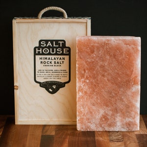 Op de afbeelding: Een grote Himalaya-steenzout kookblok in een houten doos. De doos heeft een label met de tekst "Salt House Himalayan Rock Salt Cooking Block". Het label bevat ook instructies voor het gebruik van het zoutblok.