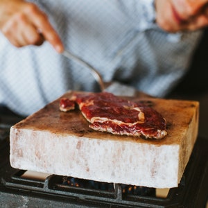 Op de afbeelding: Een rauwe steak wordt gekookt op een grote plaat roze Himalaya-zout. Het zout rust op een zwart metalen rooster.