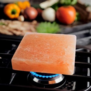 May include: A pink Himalayan salt block, rectangular in shape, is positioned above a lit gas burner on a black hob. The background shows fresh vegetables like tomatoes, onions, and peppers, indicating a cooking context.