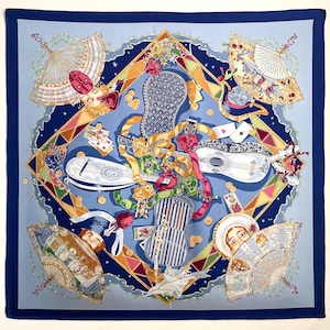 May include: A square silk scarf featuring a light blue background with a dark blue border. The design includes four decorative fans, three stringed instruments, playing cards, and gold coins arranged in a diamond pattern with ribbons.