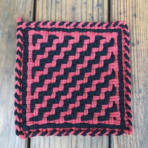 May include: A red and black woven pot holder with a braided border. The pot holder is made of cotton yarn and has a geometric pattern.