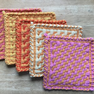 May include: A collection of five handmade woven potholders in various colors. The potholders feature a mix of pink, orange, yellow, and white hues. Each potholder has a unique woven pattern and a loop for hanging. The potholders are arranged on a wooden surface.