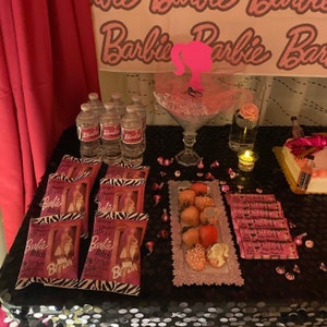 May include: A party table decorated with a Barbie theme. It features water bottles, snack bags, chocolate-covered strawberries, Hershey bars, and a cake. A banner with the word "Barbie" is visible.