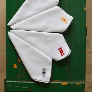May include: Four white handkerchiefs with embroidered designs in blue, yellow, red, and grey. The handkerchiefs are folded and arranged in a fan shape on a green wooden surface.
