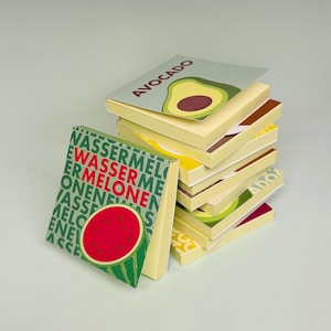 Könnte beinhalten: Ein Stapel quadratischer Haftnotizblöcke mit verschiedenen Fruchtdesigns. Der oberste Block zeigt eine Avocado-Illustration mit dem Text AVOCADO. Ein weiterer Block zeigt eine Wassermelonen-Grafik mit dem wiederholten Text WASSERMELONE in Grün und Rot.