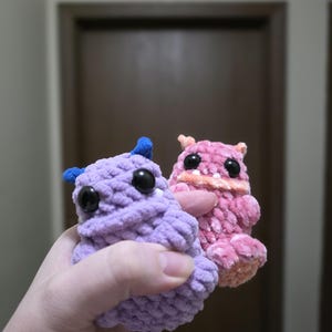 May include: Two handmade crochet creatures. One is lavender with blue horns, the other is pink with orange accents. Both have large black eyes. The plush toys are soft and cuddly, perfect for children or collectors.