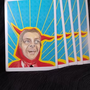 Peut inclure: Plusieurs impressions d'art représentant un portrait d'une personne dans un costume à capuche rouge avec des cornes jaunes. Le fond est bleu avec des rayons jaunes. Les impressions sont sur du carton blanc.