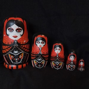 May include: A set of five nesting dolls, each painted with a red and black color scheme. The dolls feature a stylized face with red eyes and a decorative pattern. The largest doll is approximately 6 inches tall.