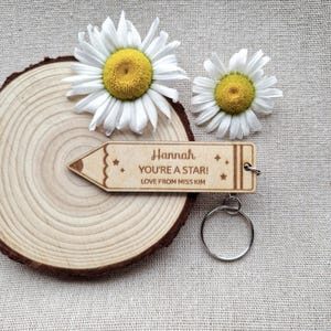 May include: A light-colored wooden keychain shaped like a pencil with the engraved text Hannah, YOU'RE A STAR!, and LOVE FROM MISS KIM, featuring small star icons. The keychain includes a silver metal ring and is positioned near two white daisies.