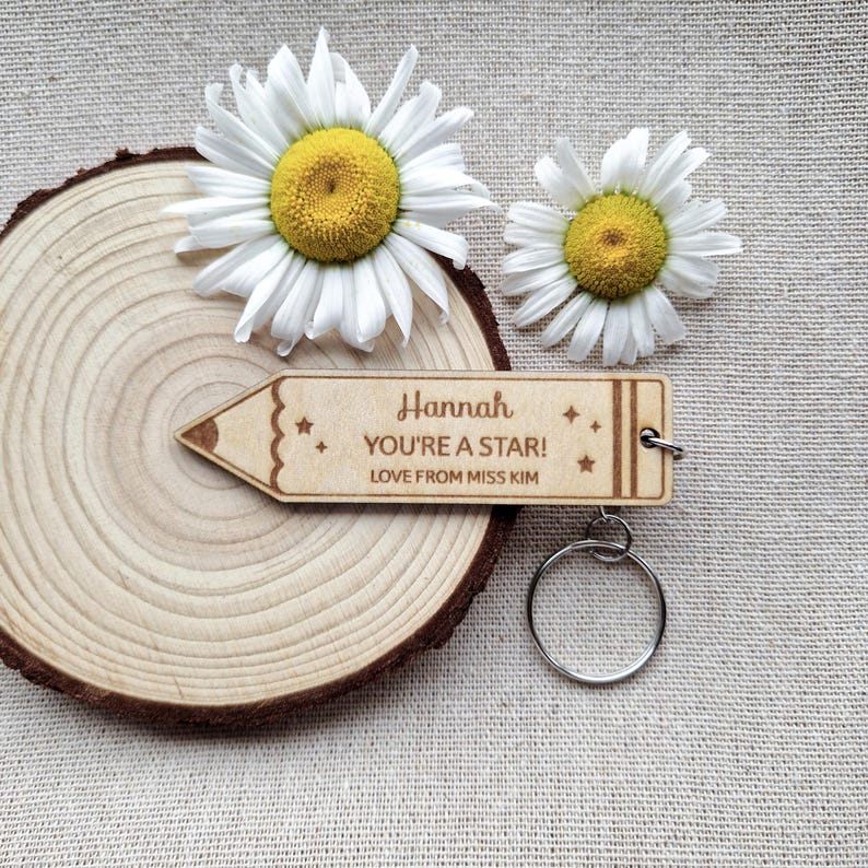 May include: A light-colored wooden keychain shaped like a pencil with the engraved text Hannah, YOU'RE A STAR!, and LOVE FROM MISS KIM, featuring small star icons. The keychain includes a silver metal ring and is positioned near two white daisies.