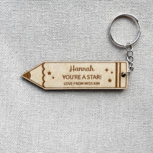 May include: A light-colored wooden keychain shaped like a pencil. The keychain features the engraved text 'Hannah', 'YOU'RE A STAR!', and 'LOVE FROM MISS KIM', along with small star icons. It includes a silver-toned metal split ring for attachment.