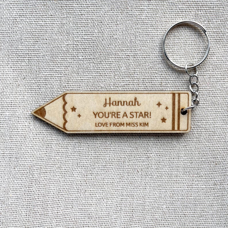 May include: A light-colored wooden keychain shaped like a pencil. The keychain features the engraved text 'Hannah', 'YOU'RE A STAR!', and 'LOVE FROM MISS KIM', along with small star icons. It includes a silver-toned metal split ring for attachment.
