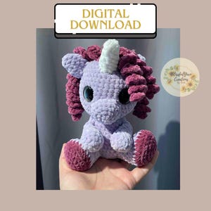May include: A crocheted purple unicorn with a white horn and pink mane. The unicorn is sitting on a hand.