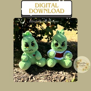 May include: Two green crocheted amigurumi toys with angry expressions. The toys are sitting on a bed of brown mulch. The toy on the left has a red heart on its chest. The toy on the right is wearing a red and white striped sweater.