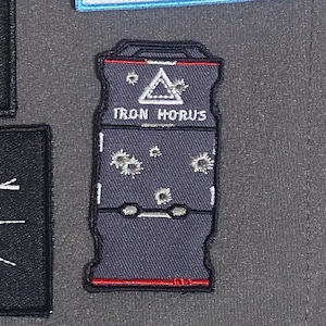 Può includere: Una toppa ricamata grigia e rossa con il testo "IRON HORUS" e un triangolo bianco con contorno nero. La toppa ha diversi fori di proiettile bianchi.