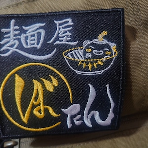 May include: Black embroidered patch with white and yellow lettering. The patch features the Japanese characters "麺屋" (menya) and "豚" (buta) and a cartoon image of a bowl of ramen with a pig face.