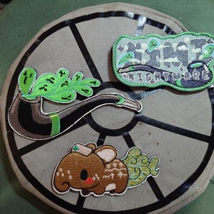 May include: Embroidered patches featuring a pipe with green smoke, a brown cartoon animal, and a patch with the word "NIGHTMARE". The patches are on a gray circular background with black lines.