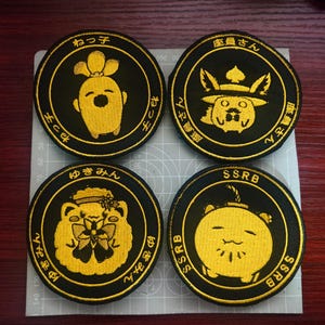 May include: Four black and gold embroidered patches with cartoon character designs. Each patch has a circular shape with Japanese text and a unique character illustration in the center. The patches are on a gray surface.