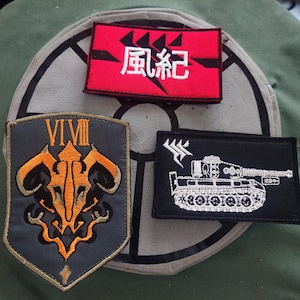 May include: Three embroidered patches with military-inspired designs. One red patch with white Japanese characters, a black patch with a tank illustration, and a shield-shaped patch with orange and gold detailing.