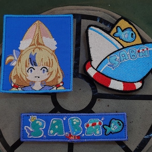 Pode incluir: Patches bordados com personagens em estilo anime e temas náuticos. Um patch retrata um personagem com cabelo loiro e orelhas de coelho em um quadrado azul. Outro mostra um barco com a palavra "SABA" e um peixe. Um terceiro patch também diz "SABA".