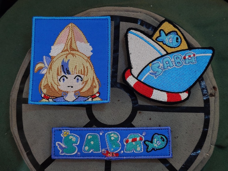 Puede incluir: Parches bordados con personajes de estilo anime y temas n&aacute;uticos. Un parche representa un personaje con cabello rubio y orejas de conejo sobre un cuadrado azul. Otro muestra un barco con la palabra "SABA" y un pez. Un tercer parche tambi&eacute;n dice "SABA".