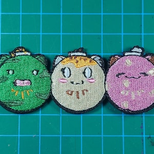 May include: Three embroidered patches with cartoon faces. The patches are round with black borders and feature green, beige, and pink colours. Each has a small hat and a unique facial expression. The patches are on a green grid background.