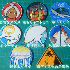 Op de afbeelding: Een verzameling geborduurde patches met diverse ontwerpen. De patches tonen stripfiguren, dieren en Japanse tekst. Kleuren zijn onder meer rood, blauw, geel en wit. De patches zijn rond en driehoekig.
