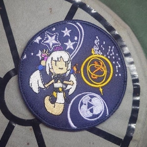 May include: Circular embroidered patch with a dark blue background, featuring a cartoon character with white hair and a black and white outfit. The design includes white stars, a clock, and gold accents. The patch is circular.