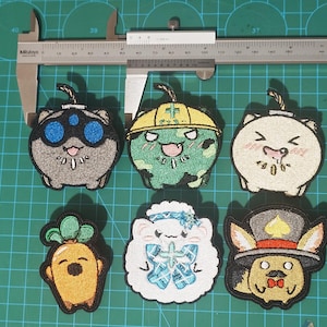 May include: Six embroidered patches featuring cartoon characters. The patches are colourful and feature a variety of designs, including a cat wearing goggles, a green creature with a yellow hard hat, a white fluffy creature with a blue bow, a yellow carrot with a face, a white cat with a pink blush, and a fox wearing a top hat.