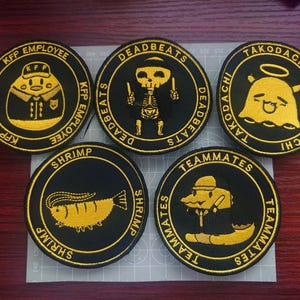 以下が含まれることがあります： さまざまなデザインとテキストが施された、黒と金の刺繍パッチが5つあります。「KFP EMPLOYEE」、「DEADBEATS」、「TAKODACHI」、「SHRIMP」、「TEAMMATES」のパッチが含まれています。各パッチは、円形のボーダー内にユニークなイラストが描かれています。