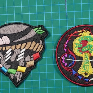 May include: Two embroidered patches on a green grid surface. One patch is a black and gray design with colorful accents. The other is a circular patch with a yellow mallet design on a black background with red and blue accents.