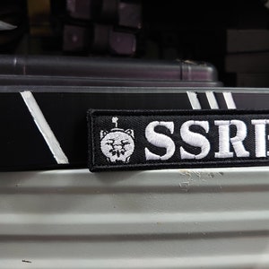 May include: A black embroidered patch with the letters SSRB in white. The patch features a cartoon character with a hat. A black box with white stripes is in the background.
