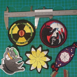 Può includere: Sei toppe ricamate con un simbolo di radiazione giallo e nero, un simbolo yin yang bianco e nero, un fiore giallo, un gatto nero, un ombrello viola e bianco e un fiocco rosso e bianco.