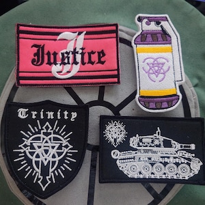Può includere: Quattro toppe ricamate con vari disegni. Una toppa recita "Justice" in corsivo bianco su sfondo rosa e nero a righe. Un'altra presenta un disegno di granata con accenti viola e gialli. Le altre due toppe presentano le parole "Trinity" e un disegno di carro armato.