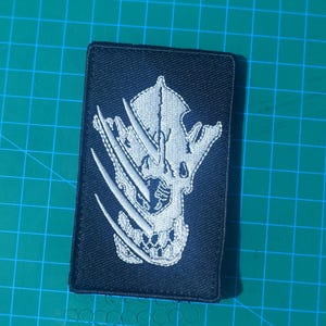 May include: A rectangular black patch with a white embroidered design. The design features a skull with three claw marks. The patch is likely for a jacket or bag.