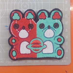 Puede incluir: Parche bordado con dos osos de dibujos animados, uno rojo y otro verde, sosteniendo un planeta rojo y blanco. Los osos tienen ojos negros y narices blancas.